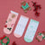 Three pairs of Christmas-themed ankle socks laid flat on a pink background with gingerbread cookies and red berries as props. The left sock is light pink with gingerbread men, stars, candy canes, and holly motifs.