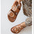 A person holds up a tan double-strap slide sandal with large gold-tone oval buckles against a light gray background, with the matching sandal also visible on the foot and a leopard-print garment shown.