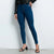 A model wearing dark blue skinny jeans, styled with a black leather belt, black leather jacket, and black strappy block-heel sandals, standing against a light gray studio background.