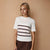 A woman modeling a white short-sleeve crewneck t-shirt with horizontal stripes in dark brown, light pink, and medium gray across the chest, set against a solid beige studio backdrop.