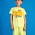 A young boy wearing a bright yellow-green t-shirt and matching shorts set, featuring a colorful graphic on the front that reads 'HELLO! SUMMER STUDY DAYS', posed against a solid blue background.