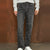 A person wearing dark gray washed denim jeans with distinctive patchwork seam details on the front of the legs, styled with a light gray sweatshirt and white sneakers against a textured brown backdrop.