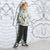 A young boy with blonde hair stands against a white brick wall with black graffiti-style drawings, wearing a pastel tie-dye hoodie, dark gray sweatpants, and black-and-white sneakers.