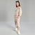 A young girl modeling a light beige two-piece lounge set consisting of a sweatshirt and matching jogger pants, paired with white sneakers, against a neutral gray studio background.