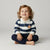 A toddler with curly light brown hair sits cross-legged on a light gray studio background, wearing a navy and white horizontally striped long-sleeve polo shirt with a small cupcake graphic on the chest, paired with navy pants.