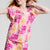 A child wearing a bright tie-dye two-piece outfit with a short-sleeve top and matching pants in pink, orange, and white, photographed against a light gray studio background.