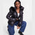 A model wearing a shiny black puffer jacket with a large black faux-fur trimmed hood, paired with blue jeans and black shoes, posing against a light gray studio background.