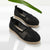 A pair of black crocheted slip-on espadrille-style shoes with natural jute-wrapped soles, photographed on a clean white surface with a green leaf prop in the background.