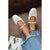 White Mary Jane flats with square toes and ankle straps, shown on a model's feet with frayed blue jeans against a brown tufted leather background.