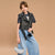A young girl modeling a black faux-leather moto jacket vest over a dark t-shirt, paired with wide-leg light blue jeans, a black studded belt, and holding a small white crescent moon handbag against a solid tan background.