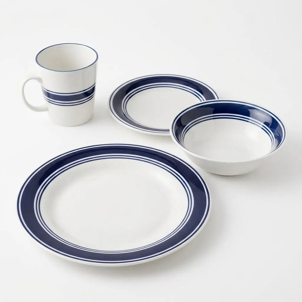 A four-piece white ceramic dinnerware set with navy blue double stripes, including a large plate, a bowl, a plate, and a mug, arranged on a clean white background.