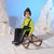 A young child wearing a bright lime green zip-up jacket, black snow pants with reflective stripes, and a dark knit beanie sits on a wooden sled in a studio set designed to look like snow with white paper mountains and a blue backdrop.