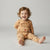 A smiling toddler sitting on a light gray studio floor wearing a tan long-sleeve pajama set with brown polka dots and a reindeer face graphic on the front.