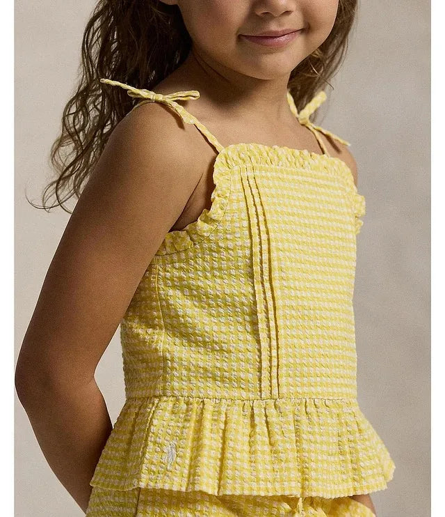 A young girl wearing a yellow and white gingham patterned sleeveless top with thin shoulder ties and a ruffled peplum hem, shown against a neutral beige background.