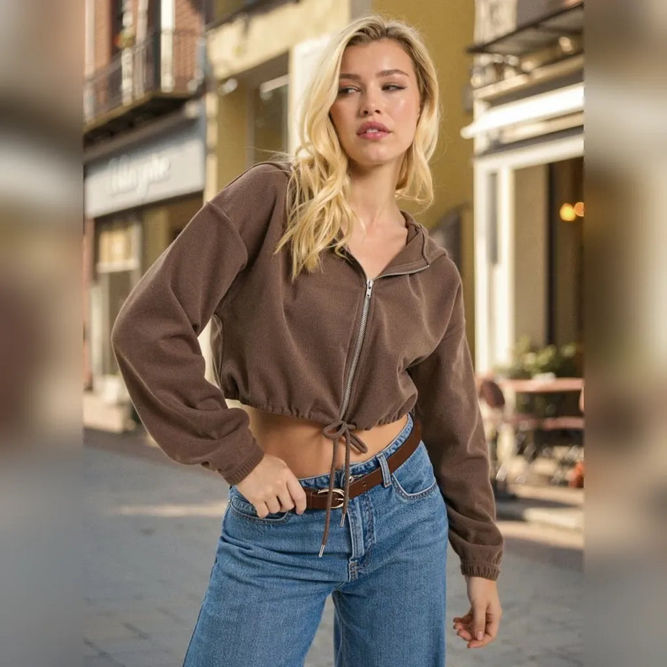 A woman wearing a brown cropped zip-up hoodie with a drawstring tie at the hem, paired with blue jeans, standing on a city street.