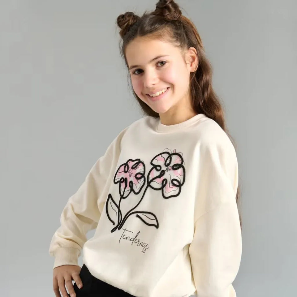 A young girl with her hair in two buns wears a cream-colored sweatshirt featuring a pink and black embroidered flower design with the word 'tenderness' written below, set against a plain gray background.