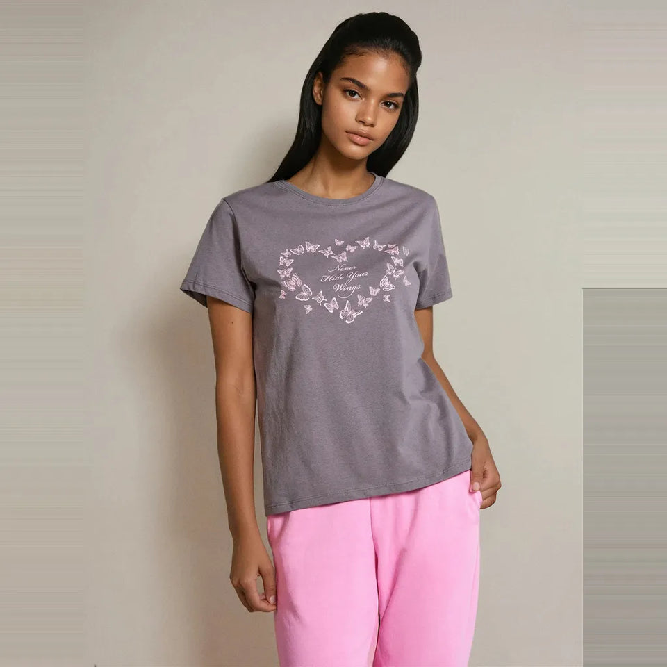 A woman modeling a heather gray short-sleeve t-shirt with a white butterfly heart graphic on the chest, paired with bright pink pants, against a neutral beige background.