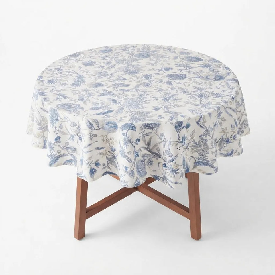 A round table covered with a white tablecloth featuring a blue floral toile pattern, supported by a wooden folding table on a clean white background.