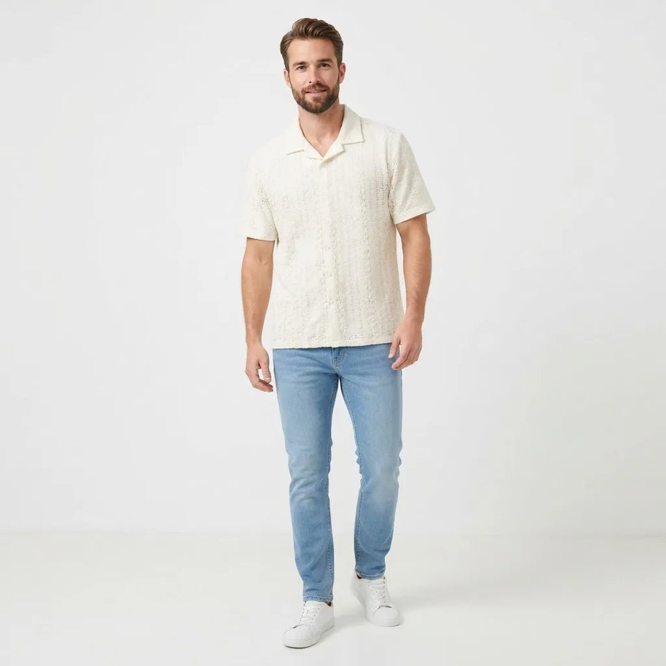 A man wearing a textured cream short-sleeve polo shirt, light blue jeans, and white sneakers, standing against a plain white studio background.