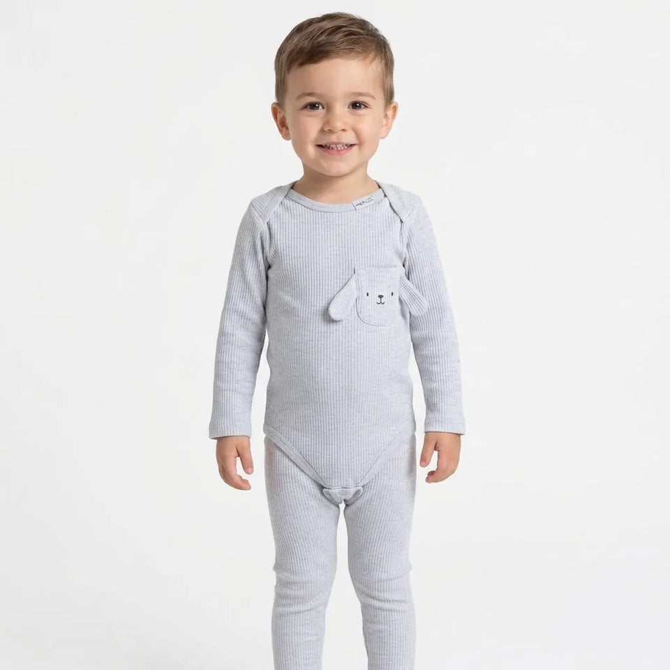 A smiling toddler stands against a plain white studio background wearing a light gray ribbed long-sleeve footed bodysuit with a small bunny face detail on the chest pocket.