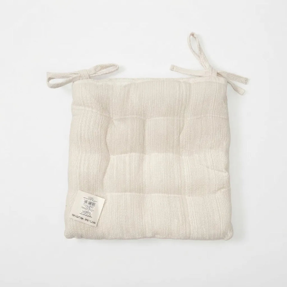 A square, quilted cotton muslin seat pad in a natural cream color, featuring tie straps at the top corners and a small sewn-in label on the lower left, shown against a plain white background.
