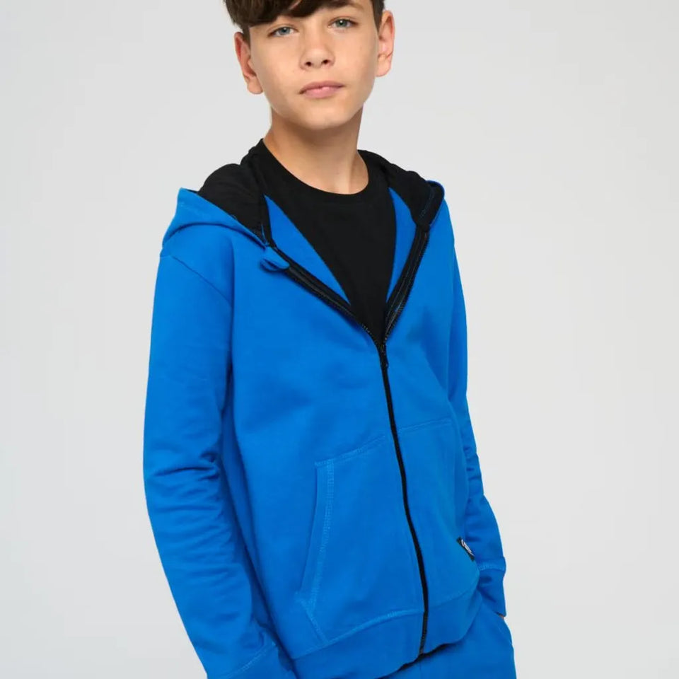 A boy wearing a bright blue zip-up hoodie with a black hood and black zipper, layered over a black t-shirt, against a light gray studio background.