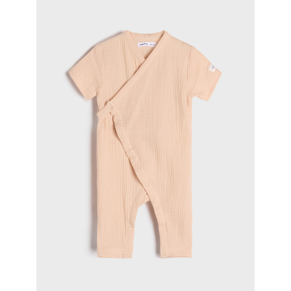 A short-sleeve baby romper in a textured, light peach-beige cotton gauze fabric, featuring a wrap-around front and leg openings, displayed flat against a light gray background.