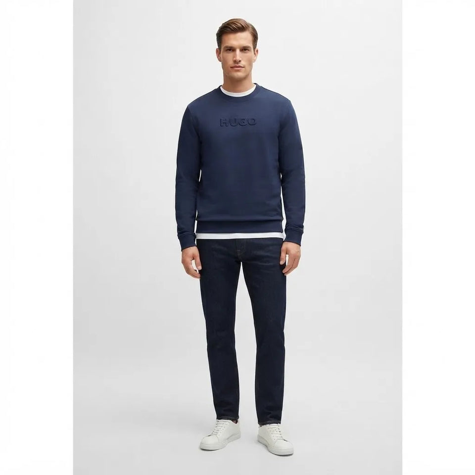 A man modeling a navy blue crewneck sweatshirt with a tonal embroidered logo on the chest, paired with dark navy pants and white sneakers, against a light gray studio background.