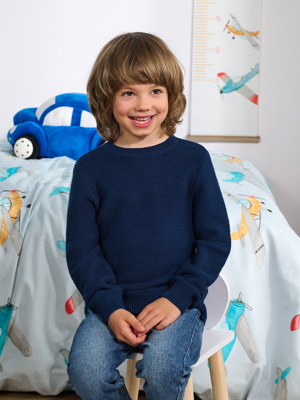 Child sitting on a chair in a room with airplane-themed bedding and toys.