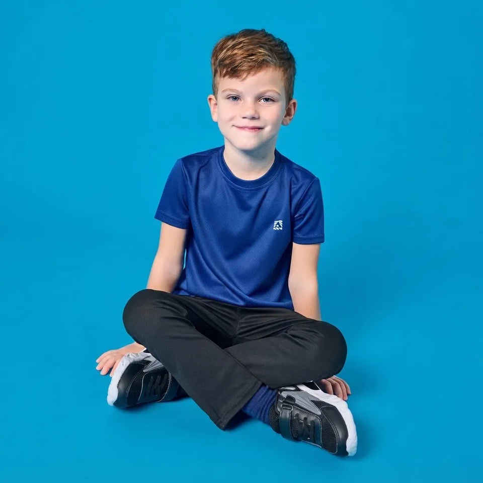 A young boy with short brown hair sits cross-legged on a solid blue studio background, wearing a royal blue athletic t-shirt with a small white logo on the left chest, black pants, and black-and-white sneakers.
