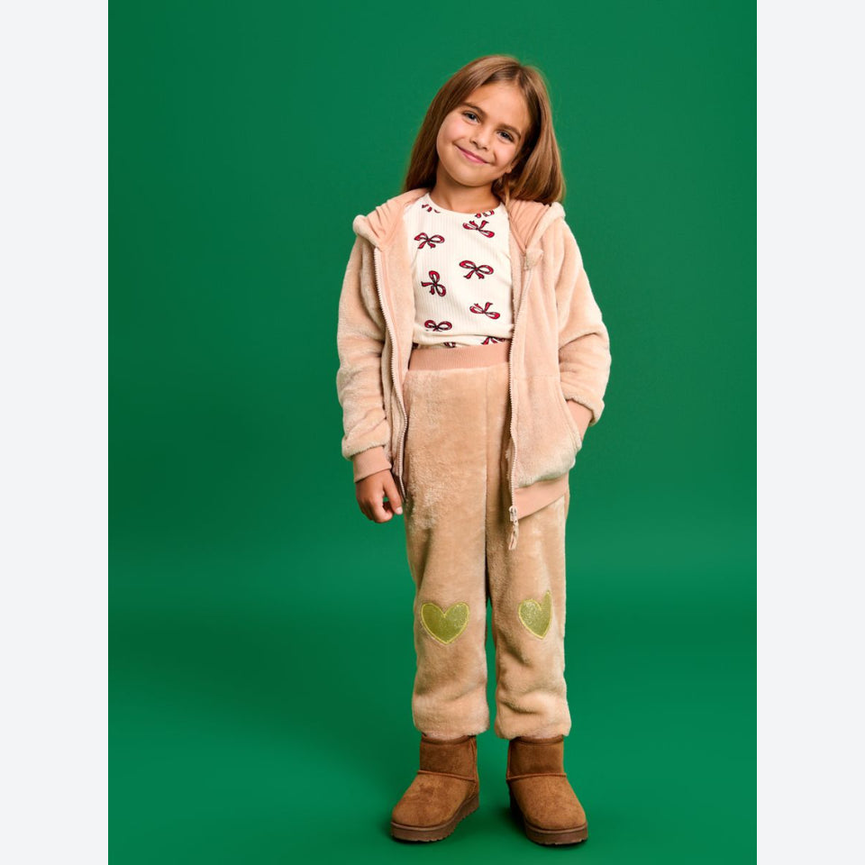 A young girl stands against a solid green studio background wearing a plush fleece matching set. The set includes a light beige zip-up hoodie with a white t-shirt underneath featuring a brown bow pattern, and matching beige pants with green heart patches.
