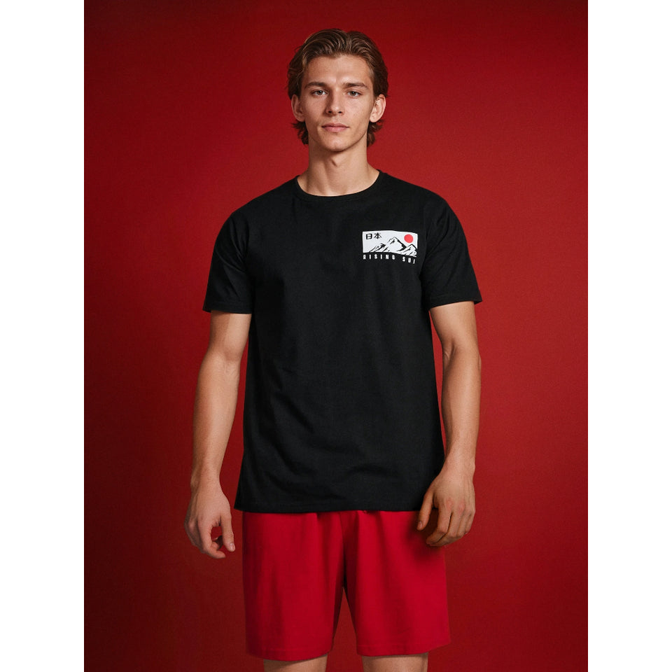 A young man modeling a black short-sleeve t-shirt with a small white rectangular chest graphic and red athletic shorts, set against a solid red studio background.