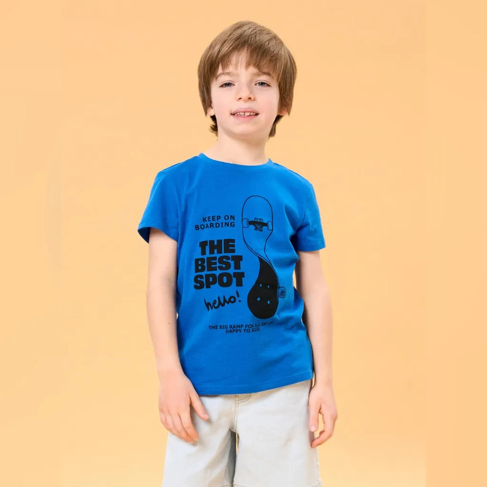A young boy with light brown hair stands against a solid warm beige background, wearing a bright blue graphic t-shirt and light beige shorts. The t-shirt features a black graphic of a fish and the text 'KEEP ON BOARDING THE BEST SPOT hello!'.