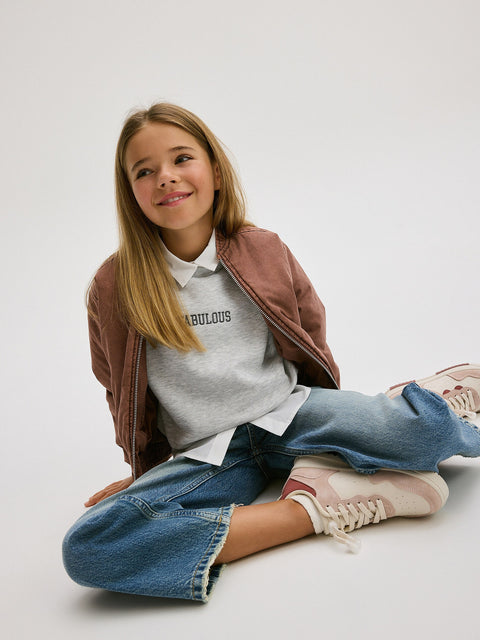 Young girl sitting on the floor wearing a brown jacket, gray sweatshirt with text, and blue jeans.