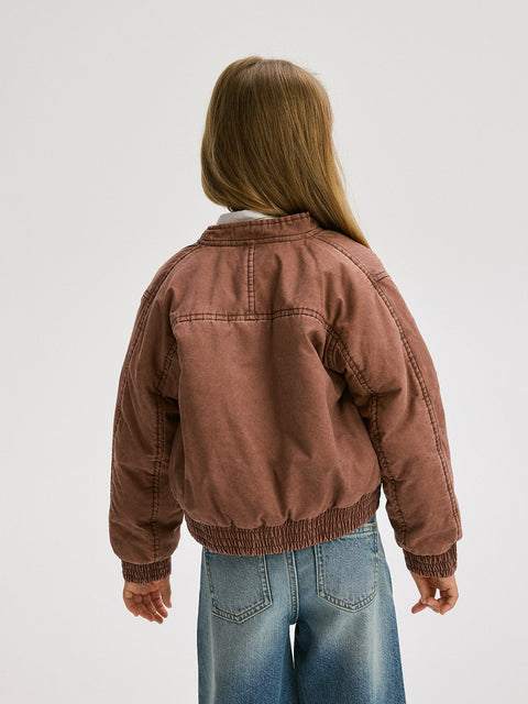 Person wearing a brown jacket and blue jeans on a plain background