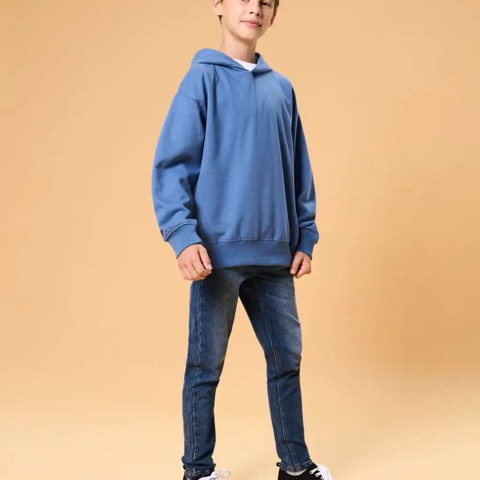 A child model wearing an oversized blue hoodie and dark blue jeans, standing against a solid warm beige background.