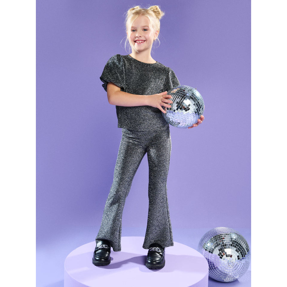 A young girl stands on a round platform wearing a sparkly dark gray short-sleeve top and matching flare pants, holding a disco ball against a lavender studio background. A second disco ball sits on the floor beside her.