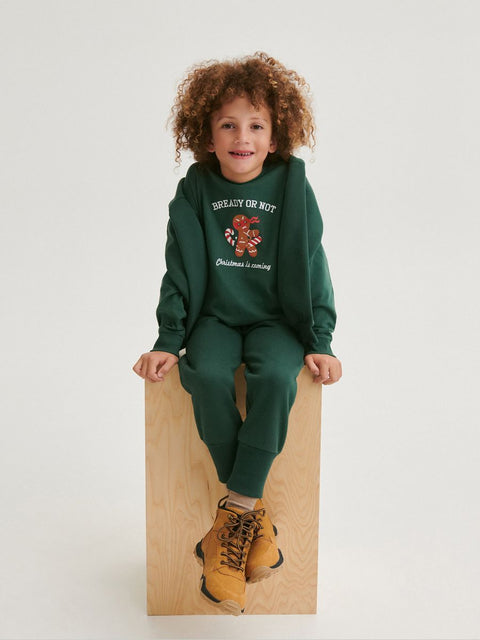 Child wearing a green hoodie and pants sitting on a wooden block against a white background