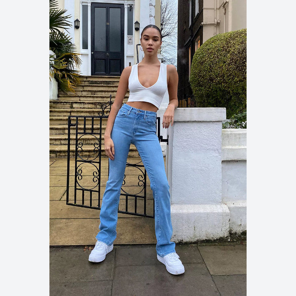 A woman stands outdoors in front of a house entrance wearing a white ribbed crop top, light blue high-waisted straight-leg jeans, and white sneakers.