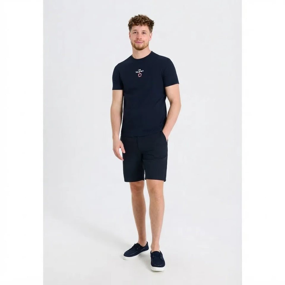 A man modeling a navy blue short-sleeve crewneck t-shirt with a small embroidered logo on the left chest, paired with matching navy shorts and dark slip-on shoes, against a light gray studio background.