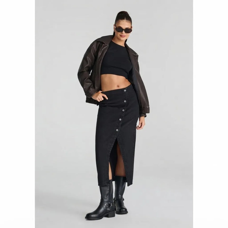 A model wearing a black denim button-front midi skirt with a front slit, styled with a black crop top, brown leather jacket, black boots, and sunglasses against a light gray studio background.