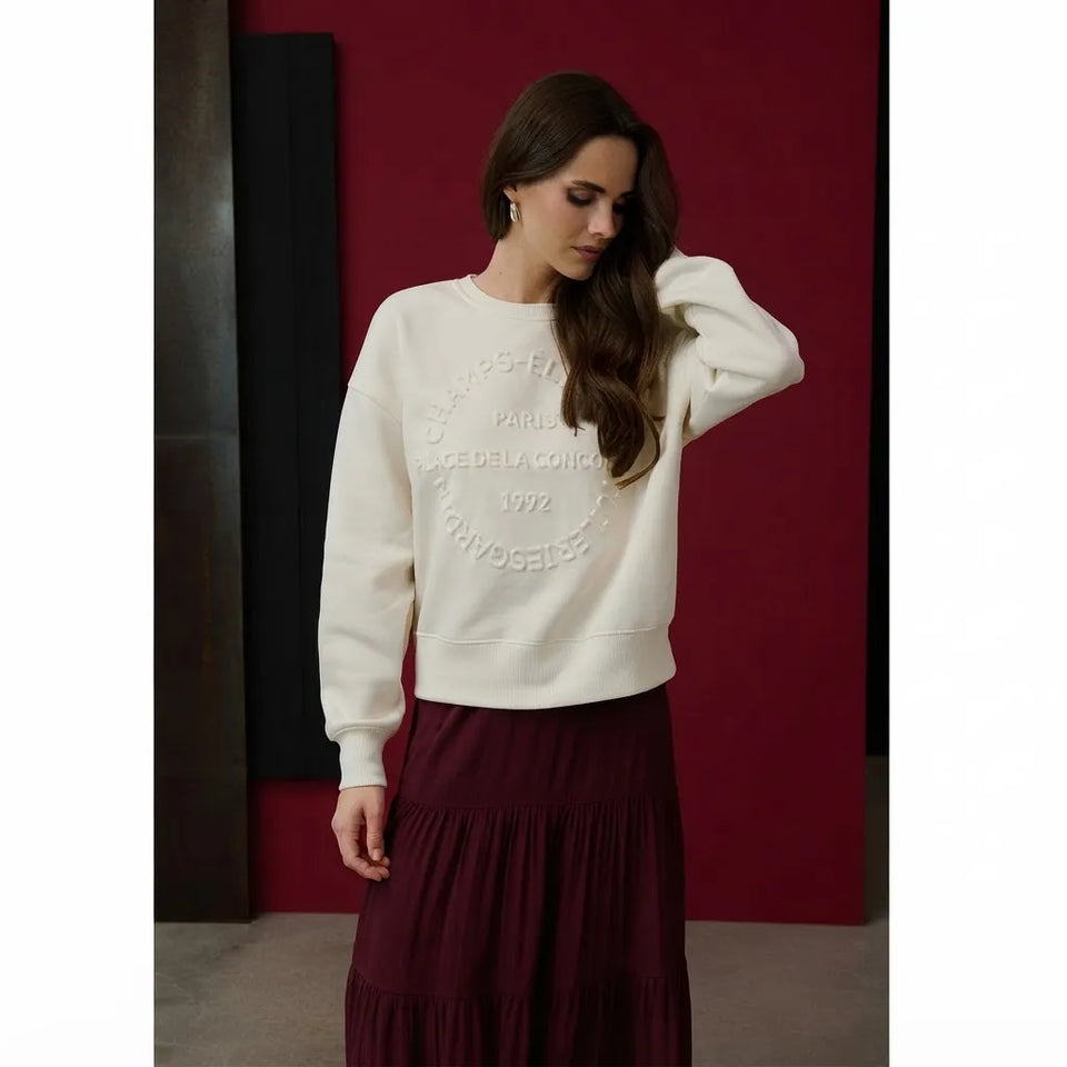 A woman wearing a cream-colored crewneck sweatshirt with tonal circular embroidery and a dark burgundy pleated maxi skirt, standing against a dark red wall.