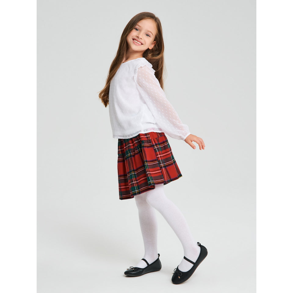 A young girl modeling a school uniform outfit consisting of a white long-sleeve blouse, a red and green plaid pleated skirt, white tights, and black Mary Jane shoes, set against a plain light gray studio background.