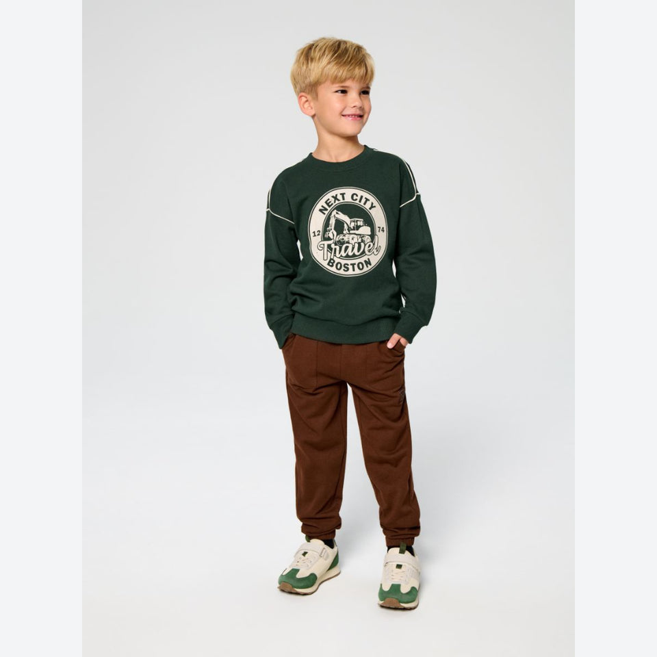 A young boy modeling a dark green crewneck sweatshirt with a white circular graphic on the chest, paired with brown pants and green and white sneakers, set against a plain light gray studio background.