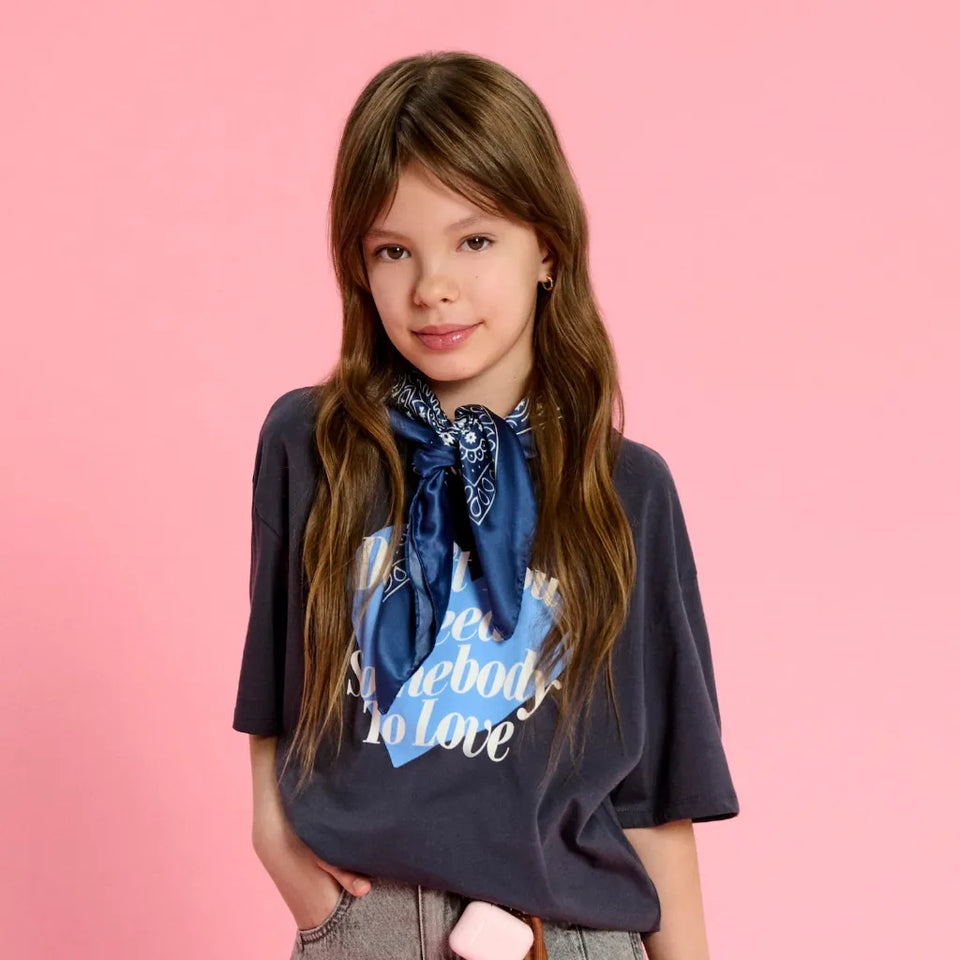 A young girl with long brown hair stands against a solid pink background, wearing a navy blue graphic t-shirt with a blue and white bandana tied around her neck.