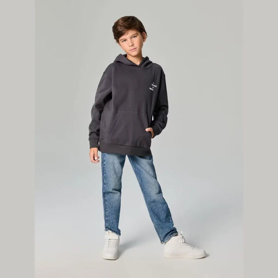 A child model wearing a dark charcoal gray hoodie with a small white graphic on the left chest, paired with blue jeans and white sneakers, standing against a light gray studio background.