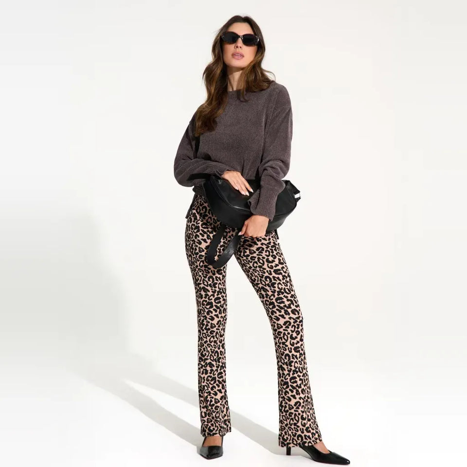 A woman modeling leopard-print flared pants with a dark brown sweater, black sunglasses, and a black crossbody bag against a plain white studio background.