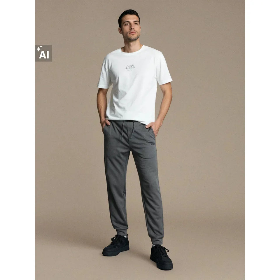 A man modeling a white crew-neck t-shirt with a small chest graphic, paired with dark gray drawstring sweatpants and black sneakers, set against a solid beige studio background.