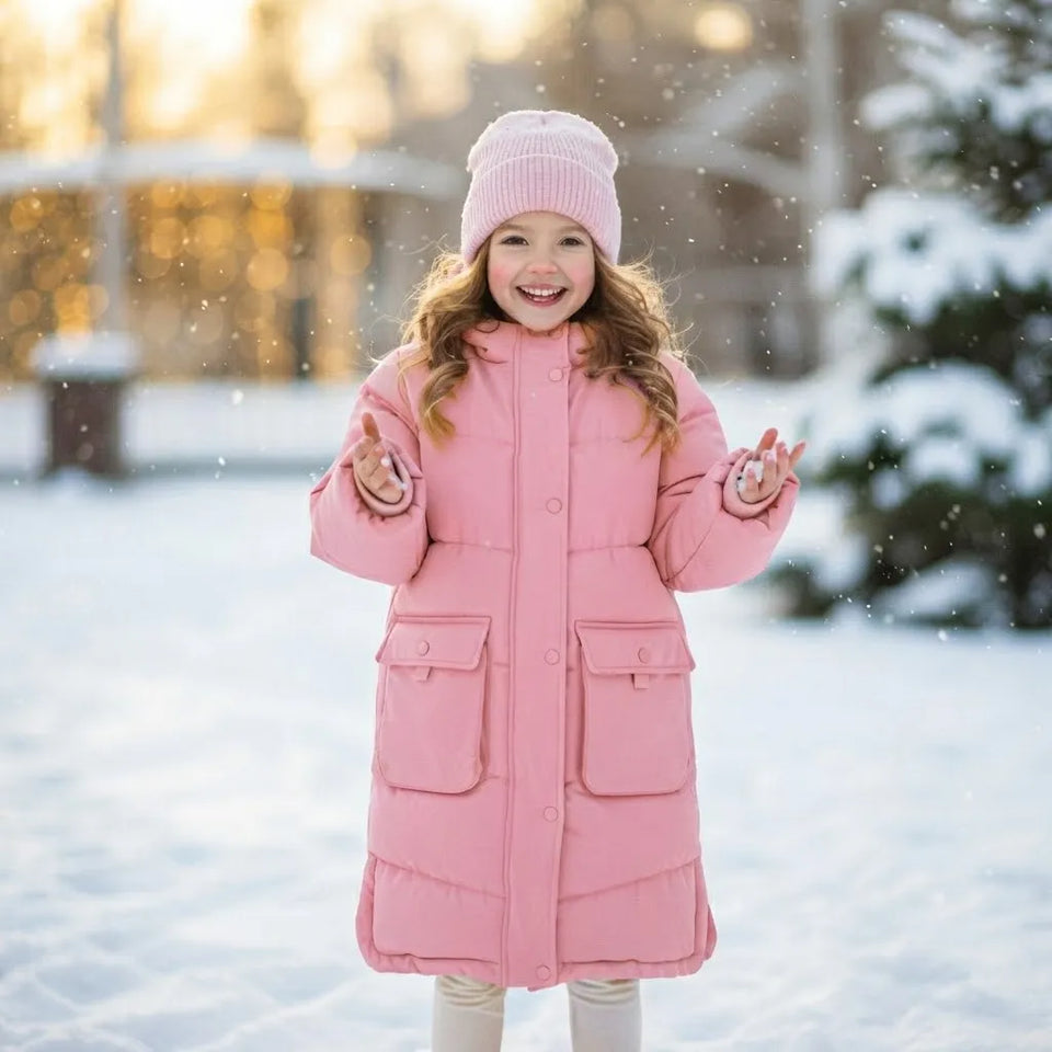 A young girl smiling in a pink puffer coat and matching pink knit beanie, standing in the snow outdoors with blurred trees and warm light in the background.