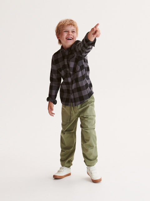 Child wearing a plaid shirt and green pants pointing upwards on a white background